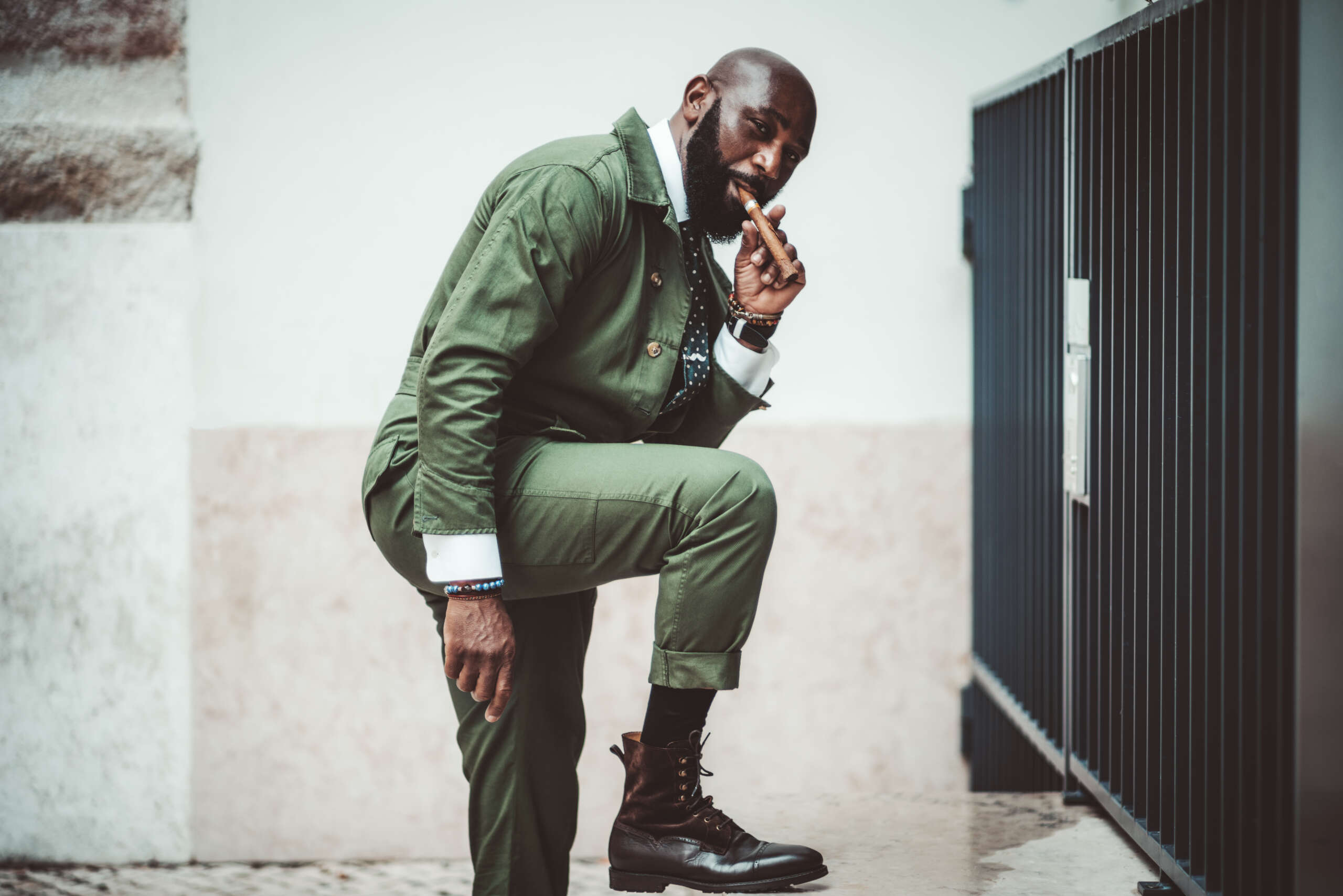 A fancy bald bearded black mature guy with a cigar and in a dark-green elegant suit and leather boots is resting his foot on a marble step in front of a wall and a fence and looking at the camera